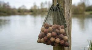 Photorealistic close-up of a dark green mesh air-dry bag filled with mixed reddish-brown and beige freezer boilies, hanging from a weathered wooden post in soft, natural daylight.
