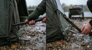 A close-up view of a heavy-duty T-peg secured into gravel ground and an adjustable tension bar keeping a carp bivvy overwrap taut in adverse weather.