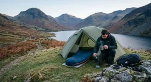 A camper kneeling outside a green tent to boil water for a hot drink, with a 4-season sleeping bag laid out inside, set against a mountainous British landscape.