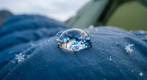 A close-up view of a water-resistant sleeping bag outer shell with tiny snowflakes resting on the fabric and a water droplet beading on the surface.